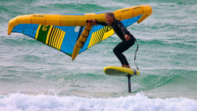 Click to go to the wing foiling blog A wing foiler rides a small wave with the wing held behind him